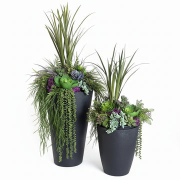 Two black planters with greenery and purple flowers on a textured surface.